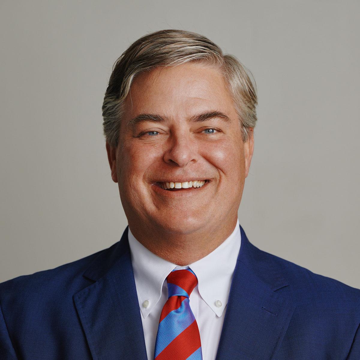 Headshot of Brent Watkins, Pancoast President. Brent wears a blue jacket and white collared shirt with a bold red and blue striped tie; he smiles wide at the camera