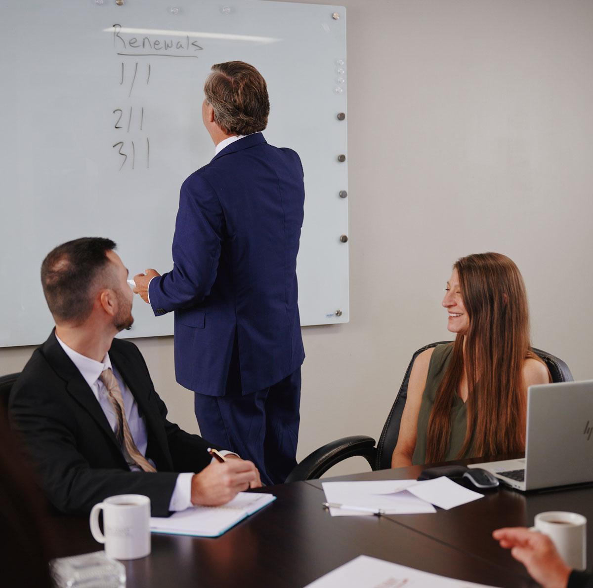 A man in a blue suit writes on a whiteboard labeled Renewals as two colleagues watch and take notes at a conference table with laptops, papers, and coffee mugs.