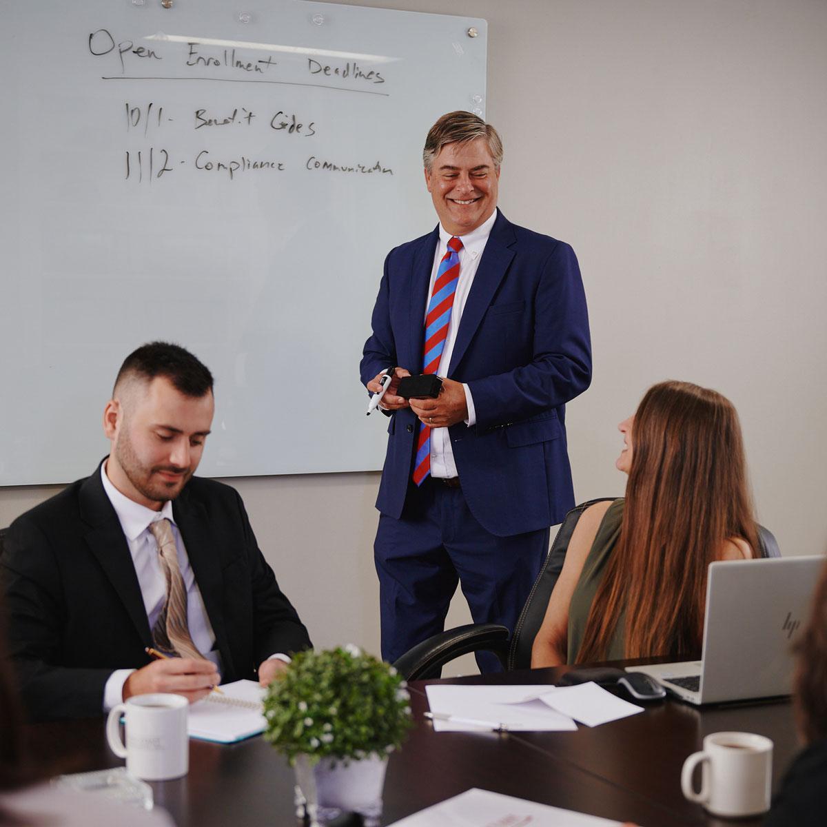 Three people in business attire meet in a conference room. One man stands smiling near a whiteboard with deadlines written on it, while two colleagues sit at a table with laptops and notes.
