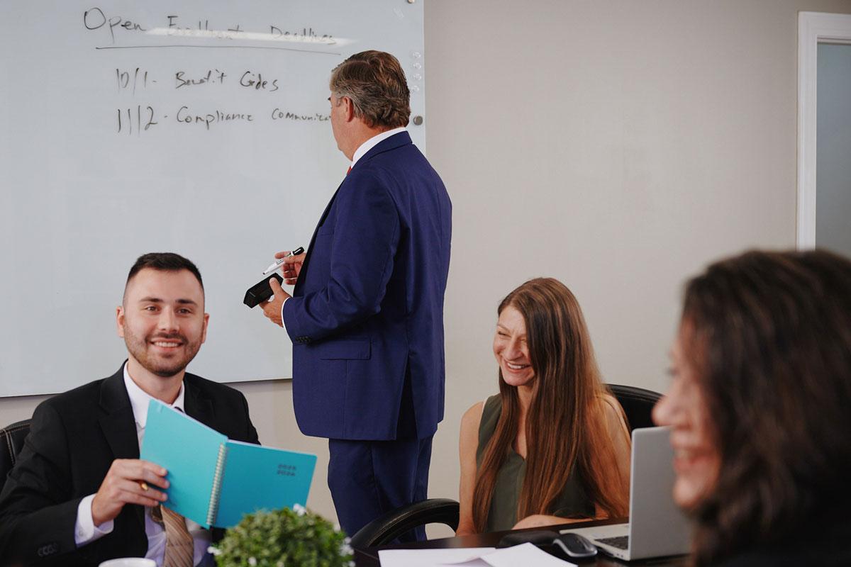 A man in a blue suit writes on a whiteboard as three coworkers sit at a conference table, smiling and talking. One man holds an open notebook, while a woman nearby laughs. A laptop and a plant are on the table.