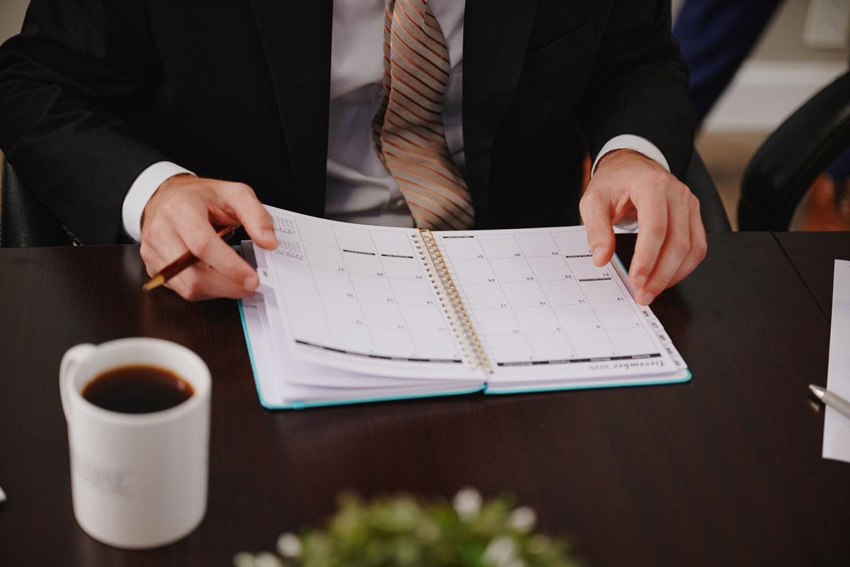 A person in a suit sits at a desk, holding a pen and looking at an open planner or calendar. There is a cup of coffee, papers, and a plant on the dark wooden table.