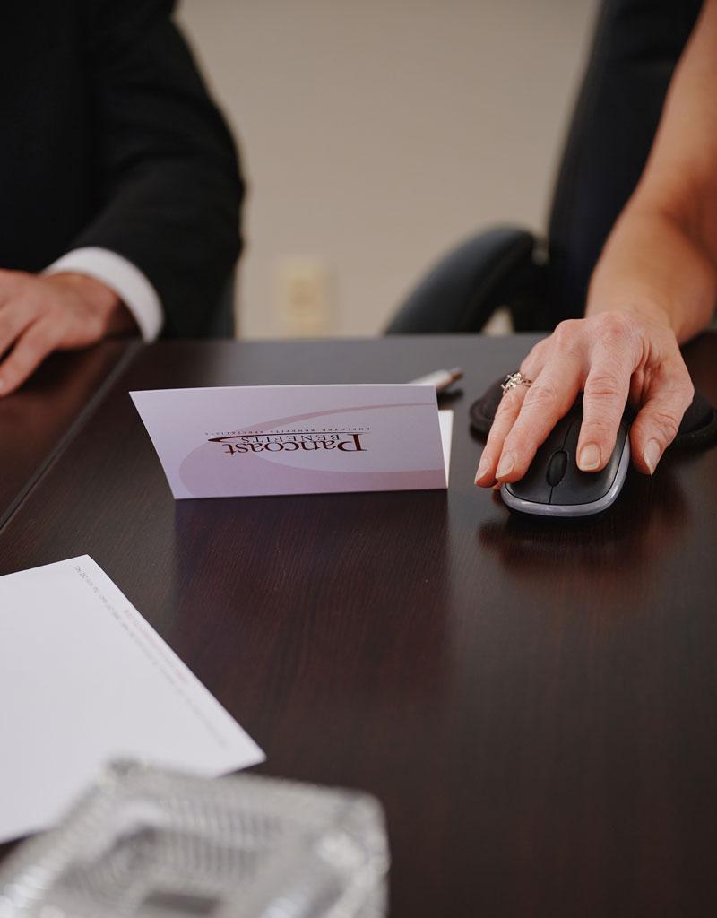 A person in business attire uses a computer mouse at a dark wooden desk, next to an envelope labeled PARDON and some papers, with another person sitting nearby.