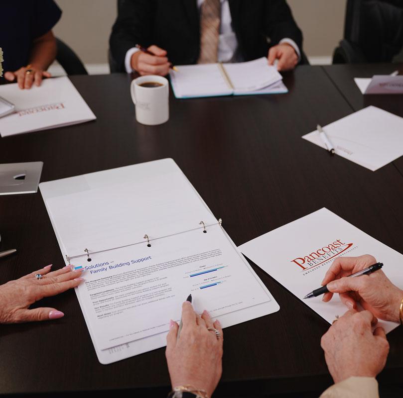 Four people sit at a conference table with documents, notebooks, a coffee mug, and a Penncorp folder. One person points at a report titled “Family Building Support,” while another takes notes.