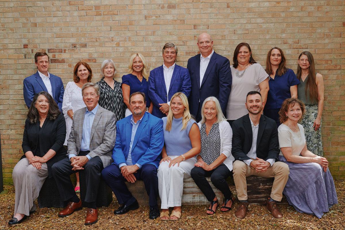 The Pancoast team - a group of 16 adults, men and women, pose together in two rows outside in front of a brick wall. They are dressed in business-casual attire and smiling at the camera.