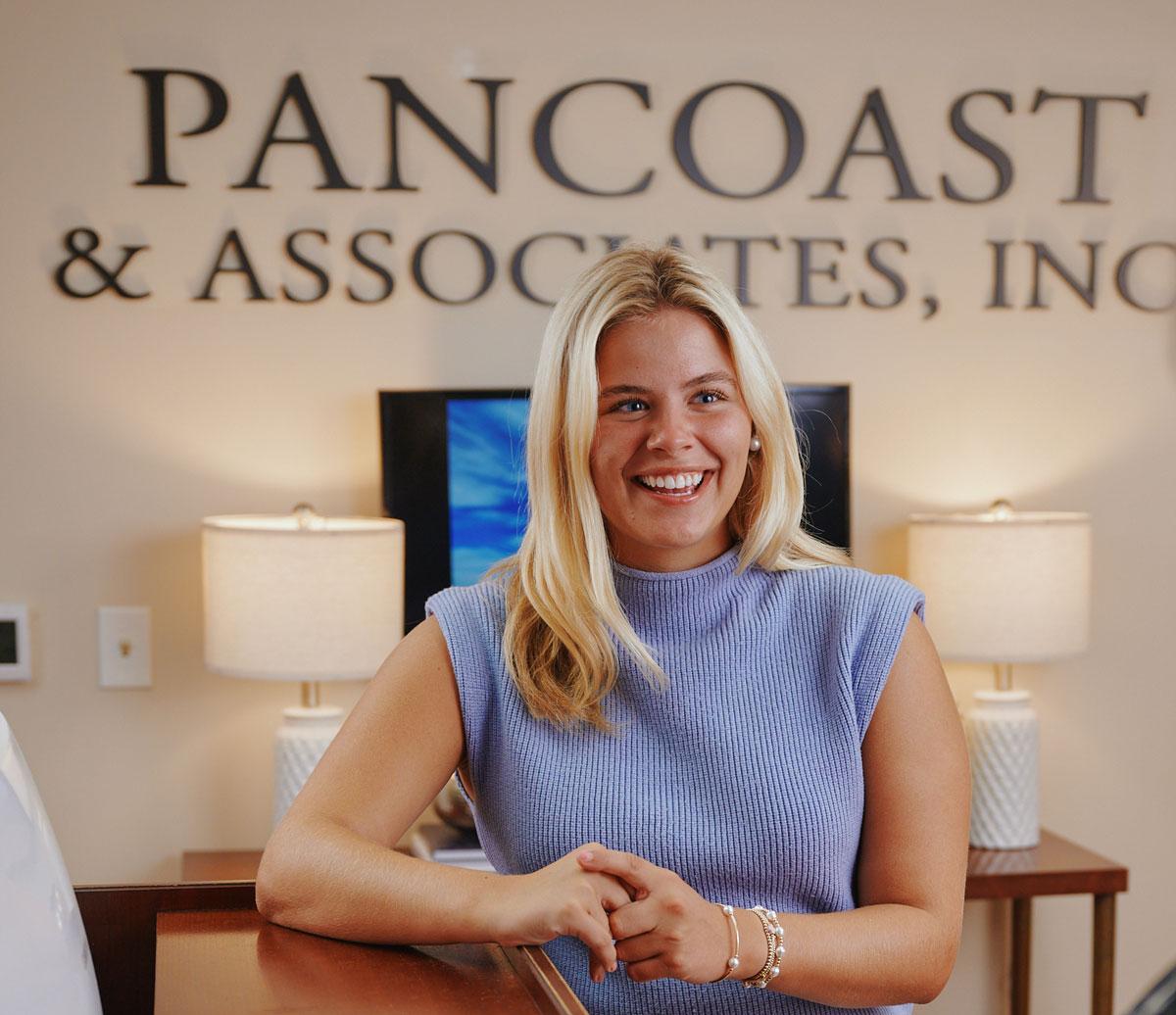 A woman with long blonde hair wearing a sleeveless blue top smiles while standing at a reception desk. Behind her is a wall sign that reads PANCOAST & ASSOCIATES, INC. and two table lamps.