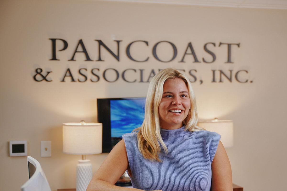 A smiling woman with blonde hair sits in an office with PANCOAST & ASSOCIATES, INC. displayed on the wall behind her, along with lamps and a TV in the background.