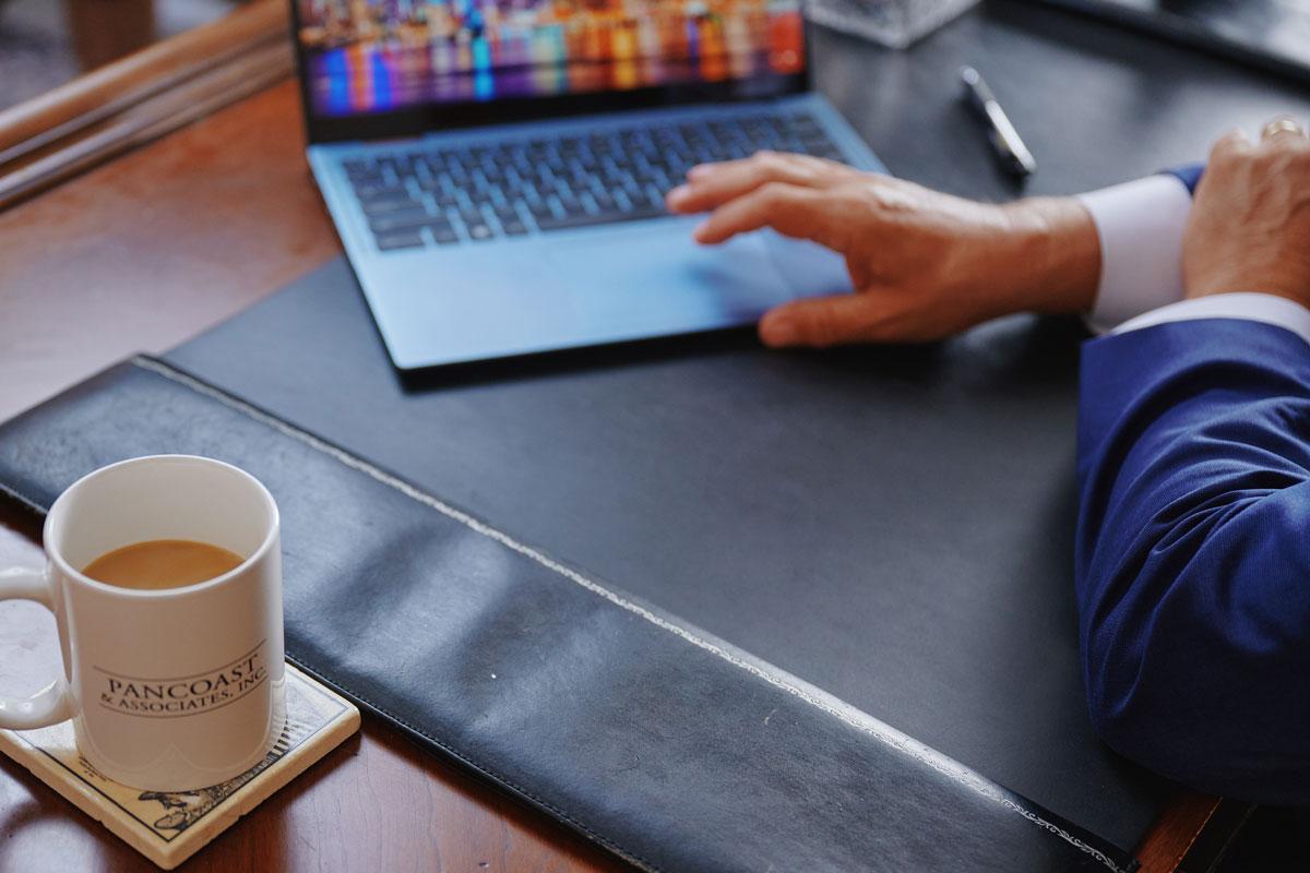 A person in a suit works at a desk with a laptop, a cup of coffee, a pen, and a coaster reading Pancoast & Associates. Only the persons hands and part of their arm are visible.
