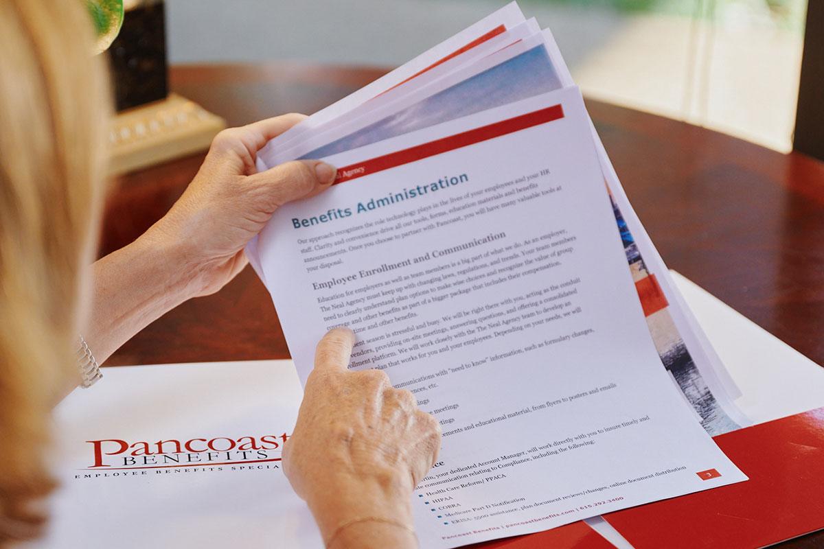 A person reads and points to a “Benefits Administration” document at a desk. A folder labeled Pancoast Benefits is partially visible underneath the papers.