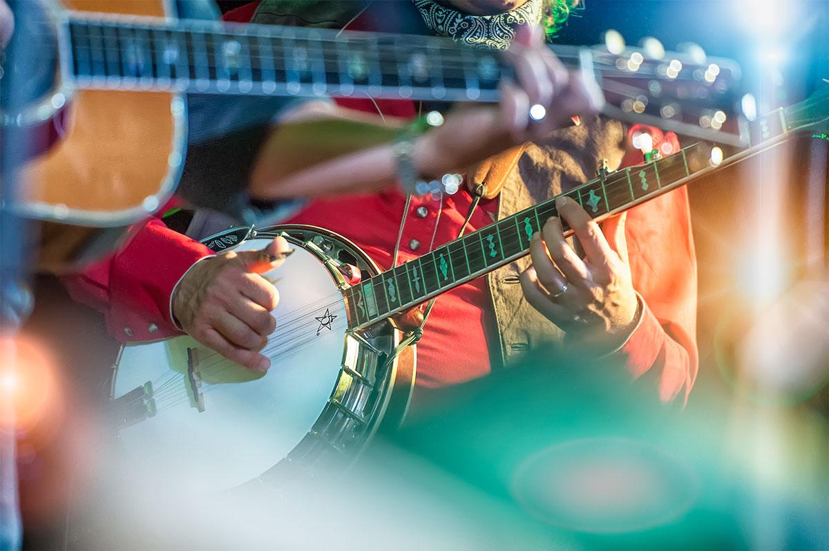Close-up of a musician in a red shirt playing a banjo, with another guitarist partially visible in the foreground. Bright stage lights and blurred colors add a vibrant, energetic atmosphere to the scene.