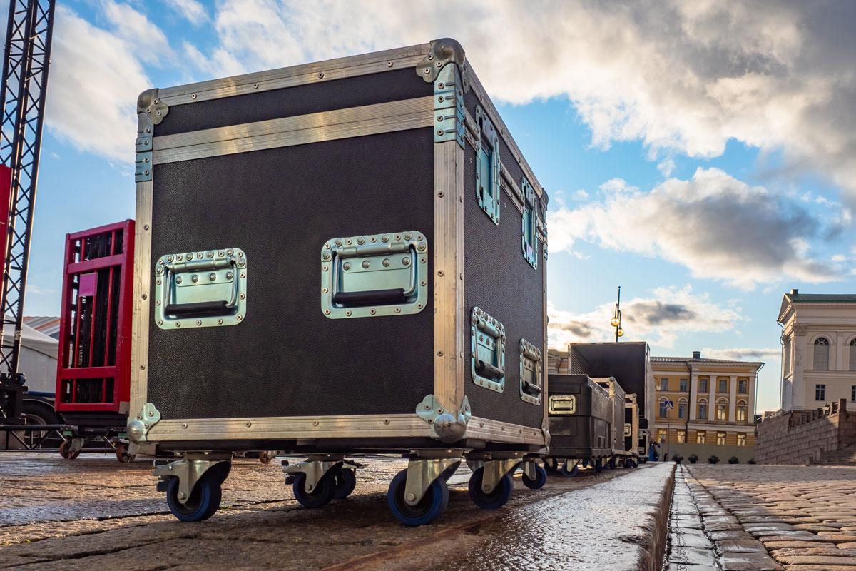 A close-up of large black equipment cases with metal edges and handles on wheels, lined up outdoors on a cobblestone surface, with buildings and a partly cloudy sky in the background.